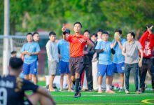Árbitro sinaliza durante uma partida de futebol, com jogadores e torcedores ao fundo.