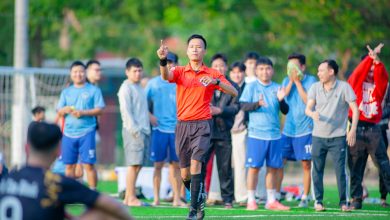Árbitro sinaliza durante uma partida de futebol, com jogadores e torcedores ao fundo.
