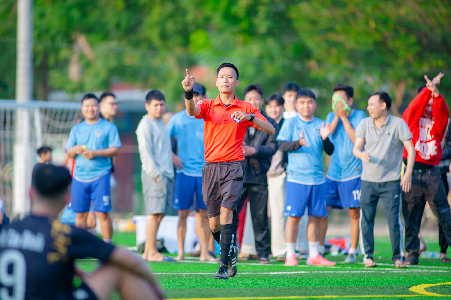 Árbitro sinaliza durante uma partida de futebol, com jogadores e torcedores ao fundo.