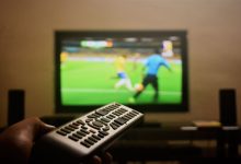 Mão segurando um controle remoto diante de uma televisão que exibe uma partida de futebol.