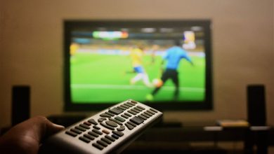 Mão segurando um controle remoto diante de uma televisão que exibe uma partida de futebol.