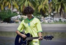 Jovem usando uma camisa amarela de futebol toca violão em uma praia tropical com palmeiras ao fundo.