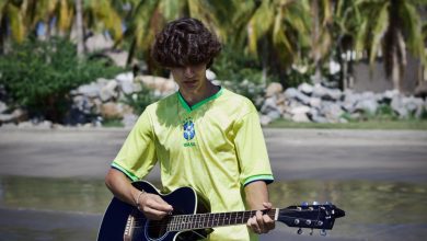 Jovem usando uma camisa amarela de futebol toca violão em uma praia tropical com palmeiras ao fundo.