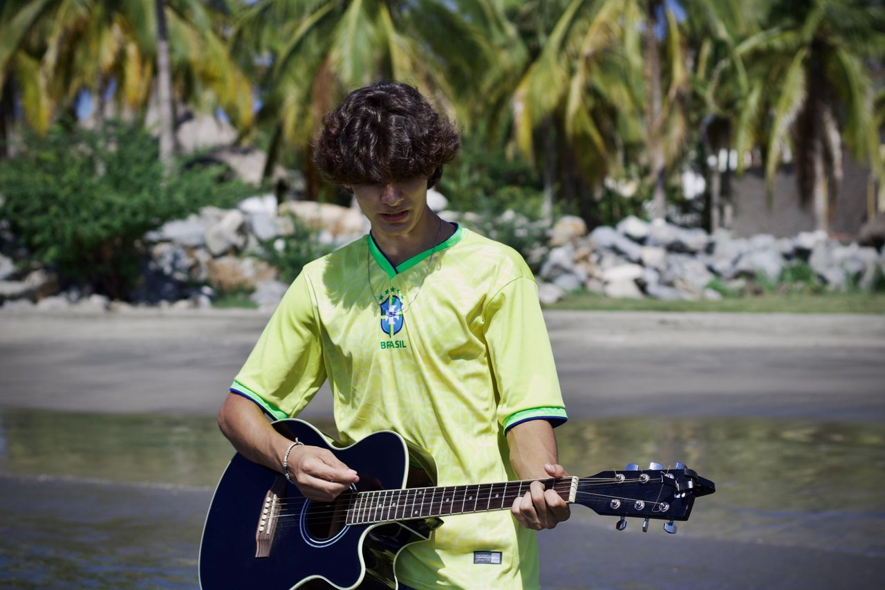 Jovem usando uma camisa amarela de futebol toca violão em uma praia tropical com palmeiras ao fundo.