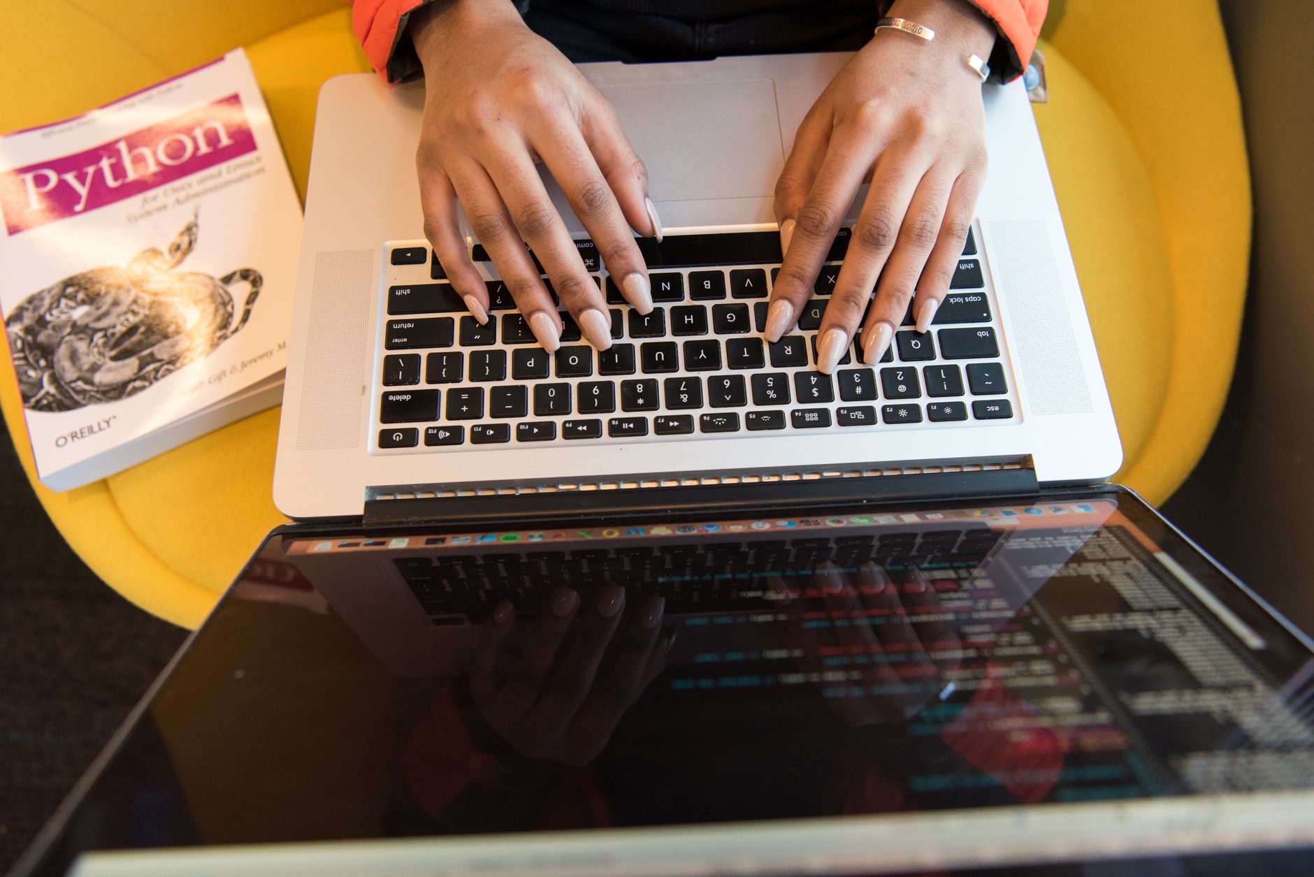 Pessoa digitando em um laptop durante uma sessão de programação, com um livro sobre Python ao lado.